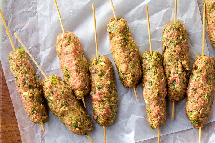 overhead shot of nine raw kofta skewers arranged on white parchment paper and a wooden surface; the skewers are made with minced meat and chopped herbs, including cilantro and green onions, they are held together by wooden skewers; the skewers are arranged in a row on the parchment paper, with the wooden skewers pointing up