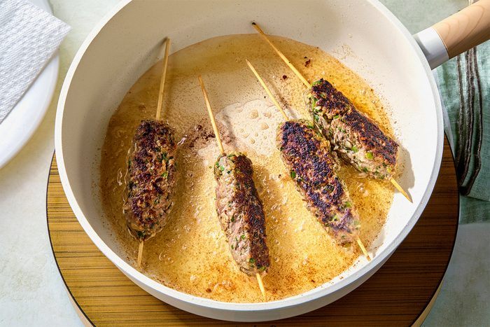 overhead shot of four kebab skewers cooking in a white pan with a wooden handle; the pan is sitting on a wooden trivet, and a white plate with a white paper towel is sitting next to it, a green and grey striped fabric is on the right; there is a lot of hot oil in the pan, and the kebabs are sizzling;