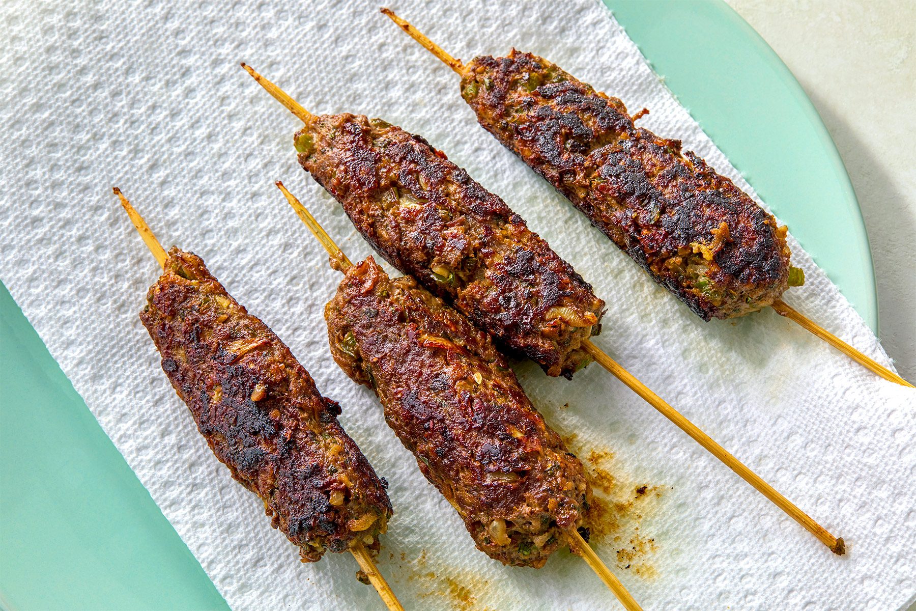 overhead shot of four kebabs on a white paper towel; the kebabs are on skewers, and they are resting on a light blue plate