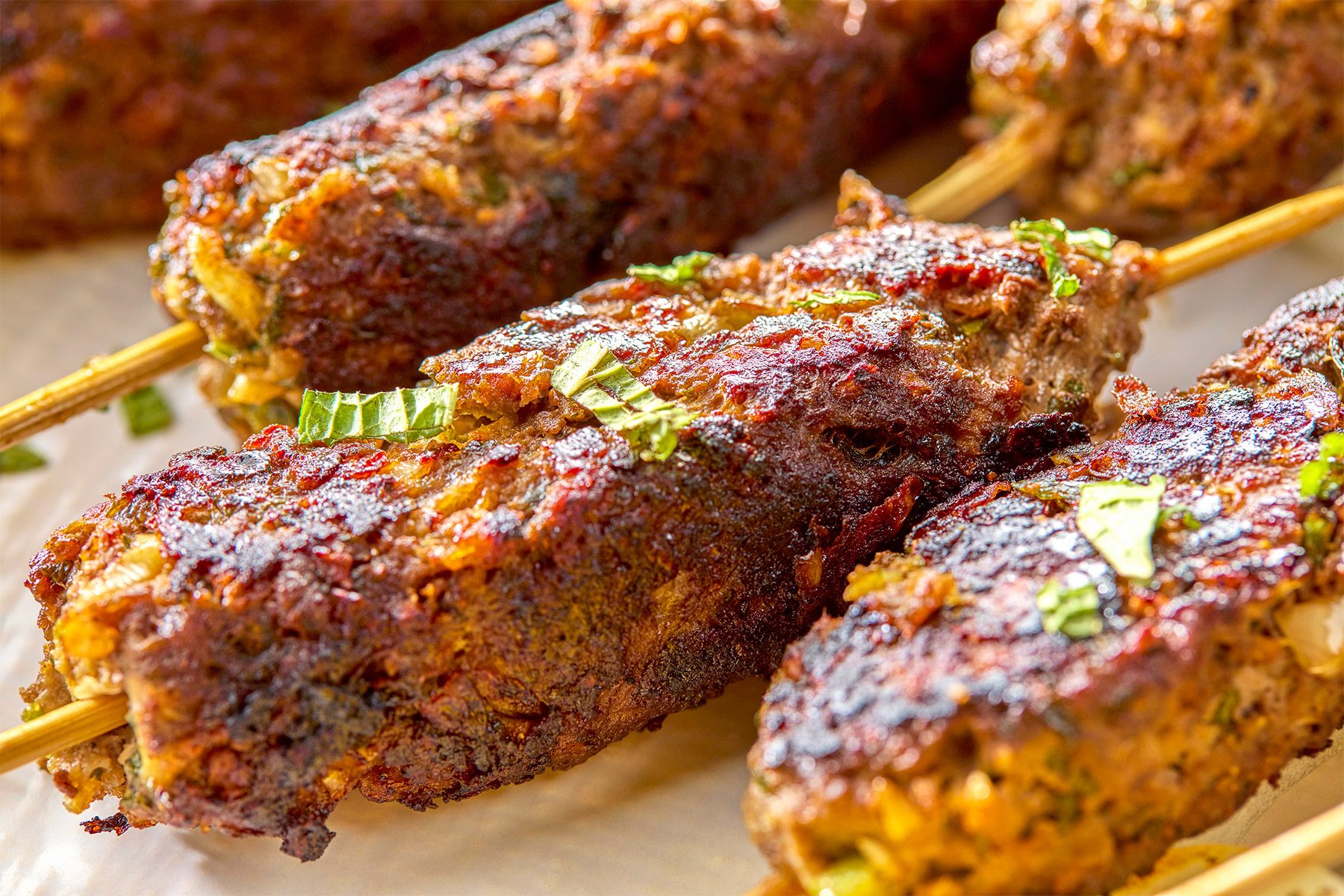 Close-up of grilled kebabs on skewers, garnished with chopped herbs. The kebabs have a crispy, browned exterior and are placed on a light-colored surface.