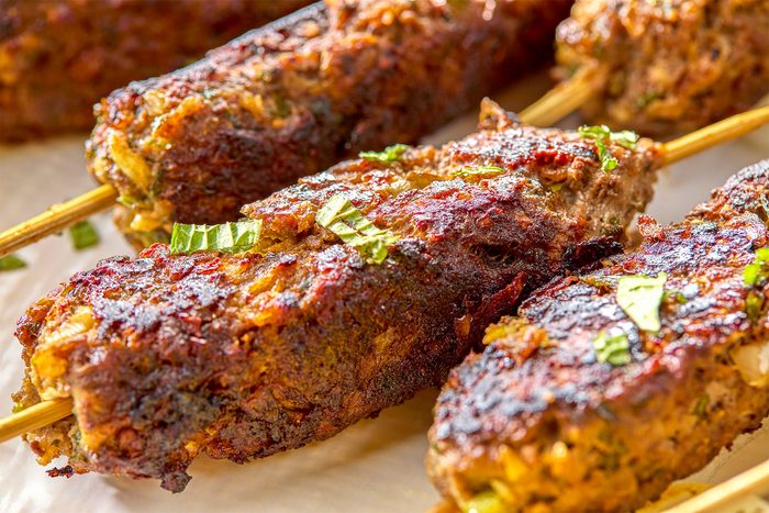Close-up of grilled kebabs on skewers, garnished with chopped herbs. The kebabs have a crispy, browned exterior and are placed on a light-colored surface.