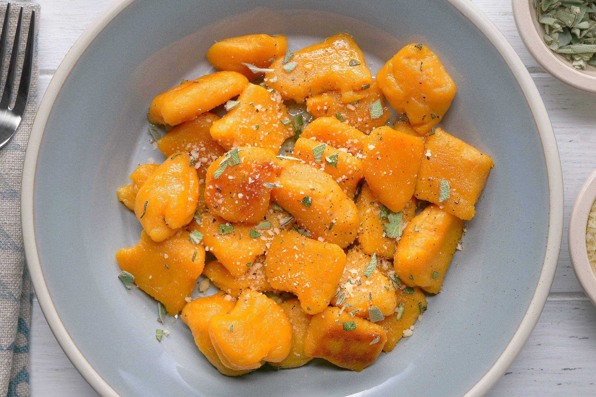 overhead shot of a plate of pumpkin gnocchi; the gnocchi is cooked and golden brown and has been tossed in a sauce of butter, sage, and parmesan cheese; the gnocchi is in a light blue bowl, and the white wood background