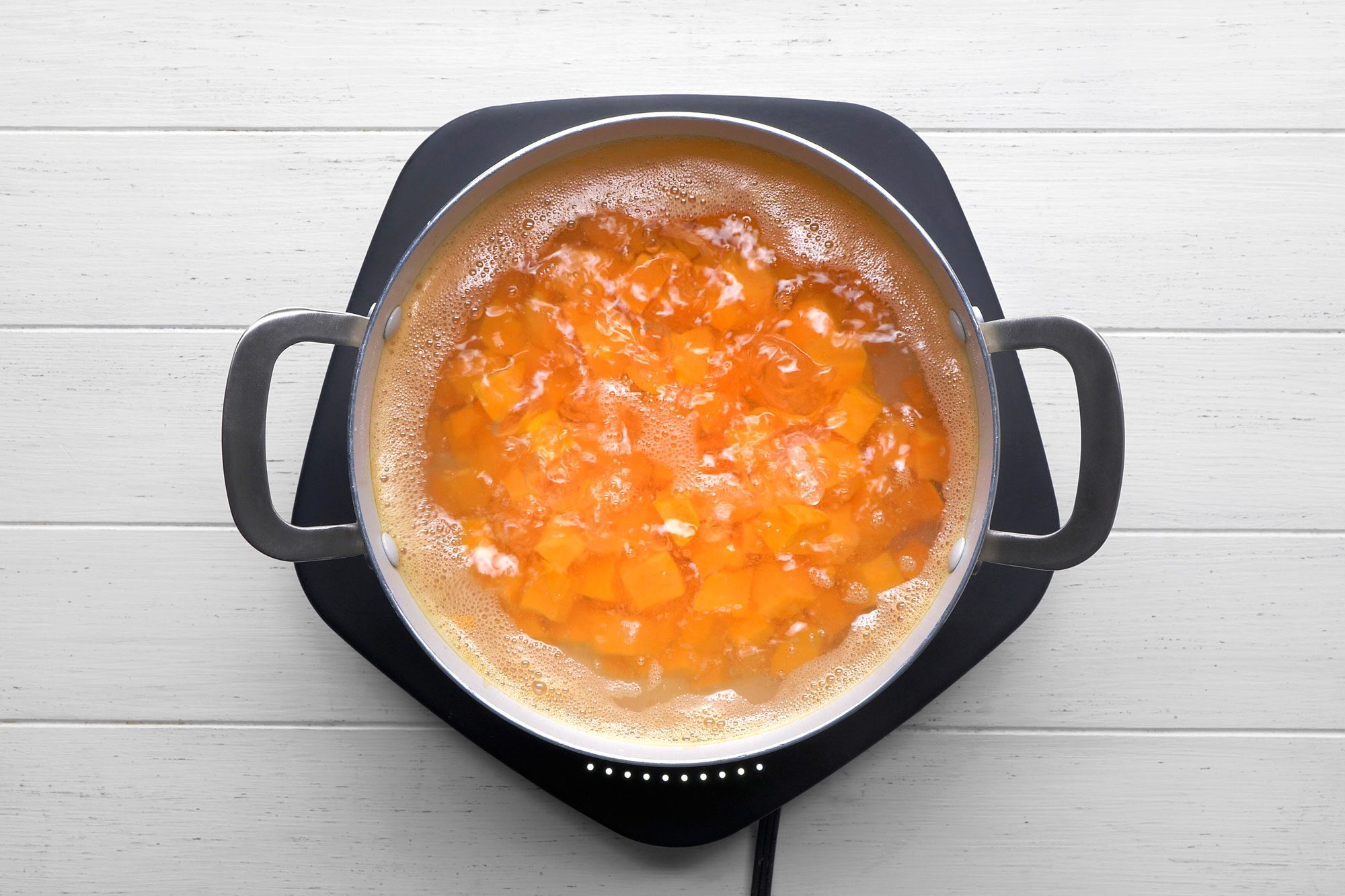 overhead shot of a pot of cubed butternut squash simmering in boiling water on a stovetop; the pot has two silver handles and the stovetop is black with white accents; the squash is a vibrant orange color and the boiling water is bubbling vigorously; the background is a white wooden surface