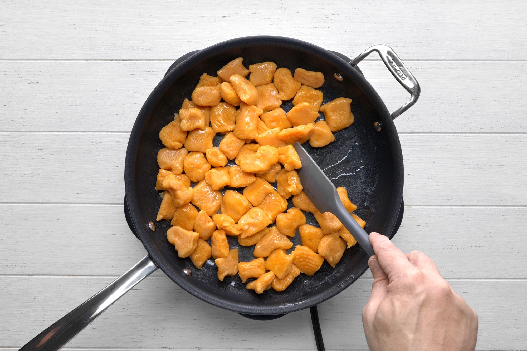 overhead shot of a black skillet with a silver handle on a white wooden surface; in the skillet, there are orange gnocchi being cooked and a hand is holding a black spatula to stir the gnocchi;