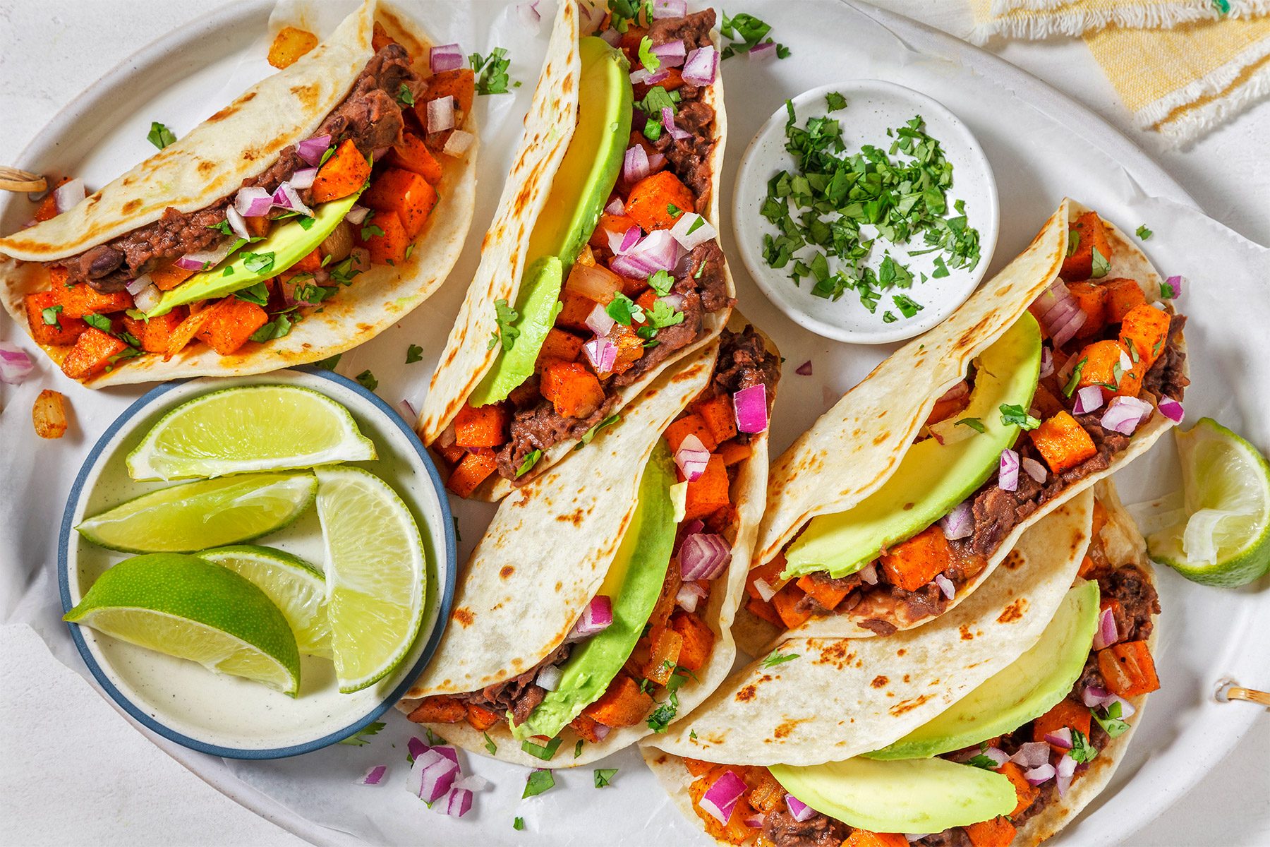 Close-up of three soft tacos on a white plate. Each taco is filled with black beans, sliced avocado, diced red onion, and roasted sweet potatoes. A garnish of fresh cilantro is visible beside the dish.