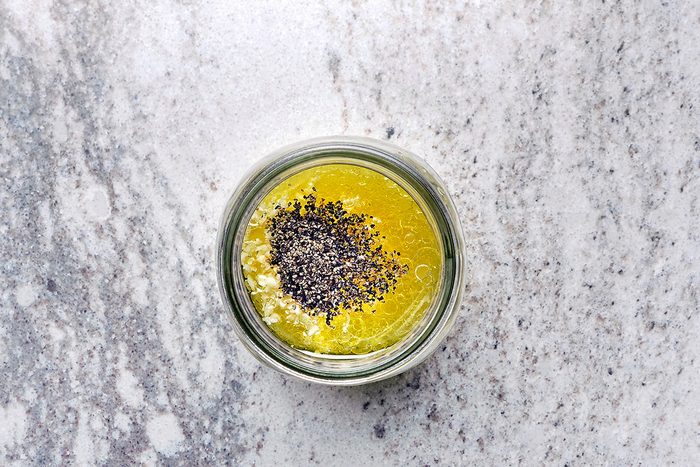 A glass jar filled with yellow oil and sprinkled black seeds on top, set on a textured gray surface.