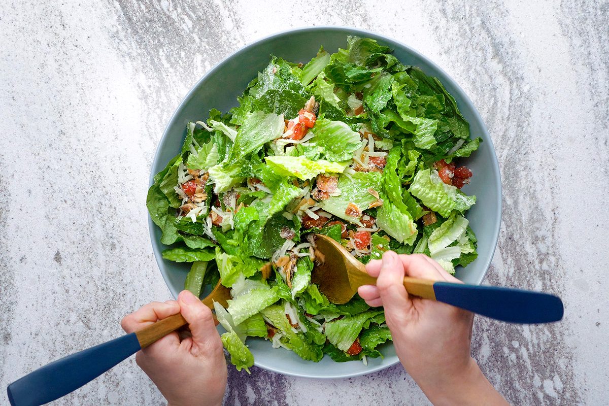 Top view of a fresh salad in a large bowl, filled with romaine lettuce, croutons, and grated cheese. Two hands are seen tossing the salad with wooden utensils on a light stone countertop.