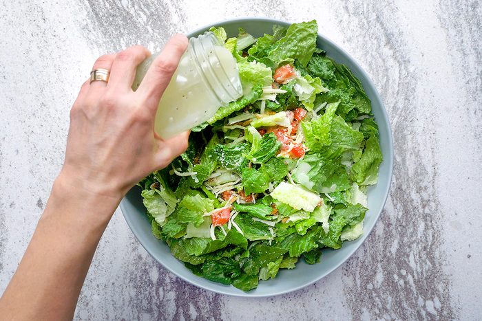 A hand pouring dressing from a jar onto a fresh salad with lettuce, tomatoes, and shredded cheese in a blue bowl. The background has a marble texture.