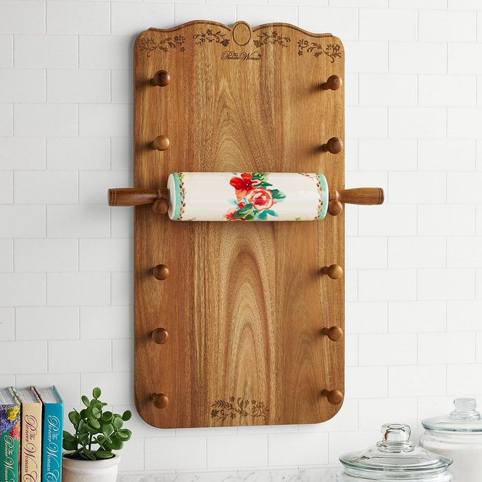 A floral ceramic rolling pin hangs on a wooden rolling pin holder with floral detailing, above books, a plant, and two jars