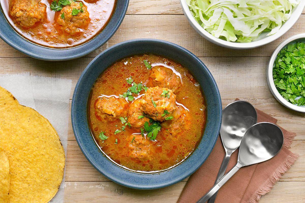 A wooden table with a bowl of spicy meatball stew, garnished with herbs. Nearby are two serving spoons, a bowl of chopped lettuce, a small bowl of cilantro, and two crispy flatbreads. Two extra bowls of stew are partially visible.