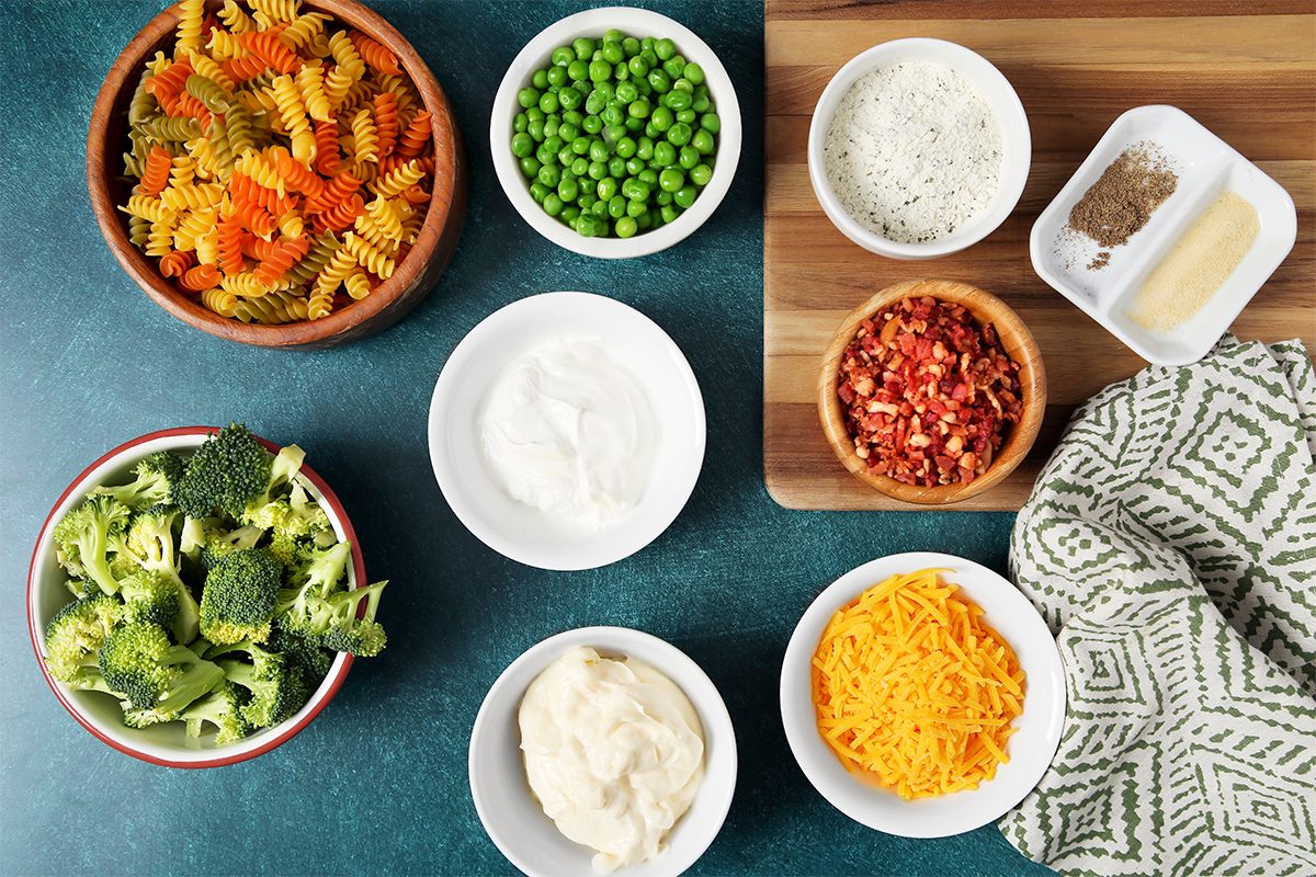 Ingredients are arranged on a table: tri-color rotini pasta, peas, broccoli, sour cream, cheese, bacon bits, grated parmesan, seasoning, and ranch dressing. A patterned cloth is nearby on a wooden board.