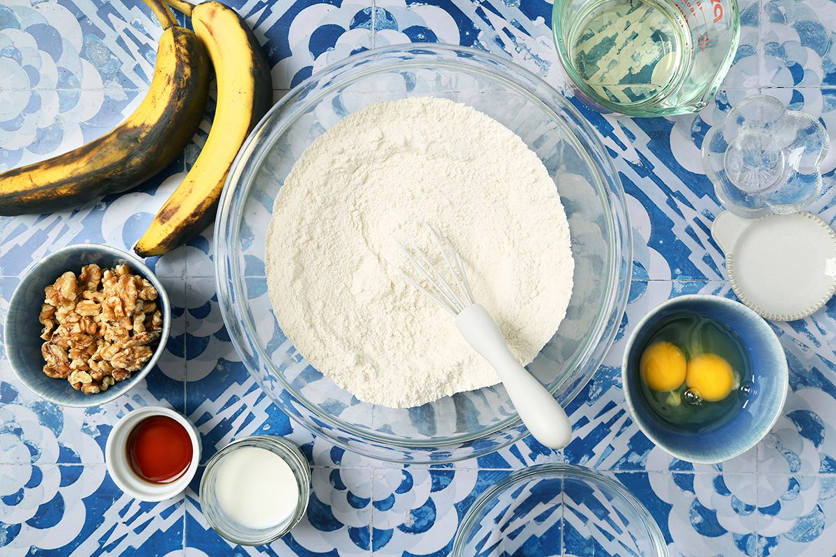 Ingredients for baking banana bread arranged on a patterned blue surface, including bananas, flour in a bowl with a whisk, eggs, milk, walnuts, vanilla extract, water, oil, and sugar.