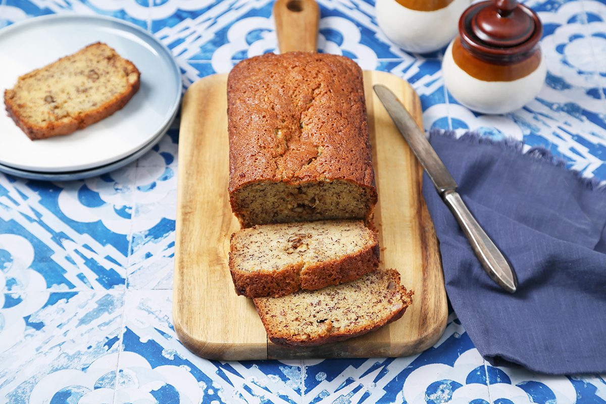 A loaf of banana bread sits on a wooden cutting board. A few slices are cut and laid out. A plate with a slice of bread, a knife, a blue napkin, and two jars are nearby, all on a blue decorative tiled surface.