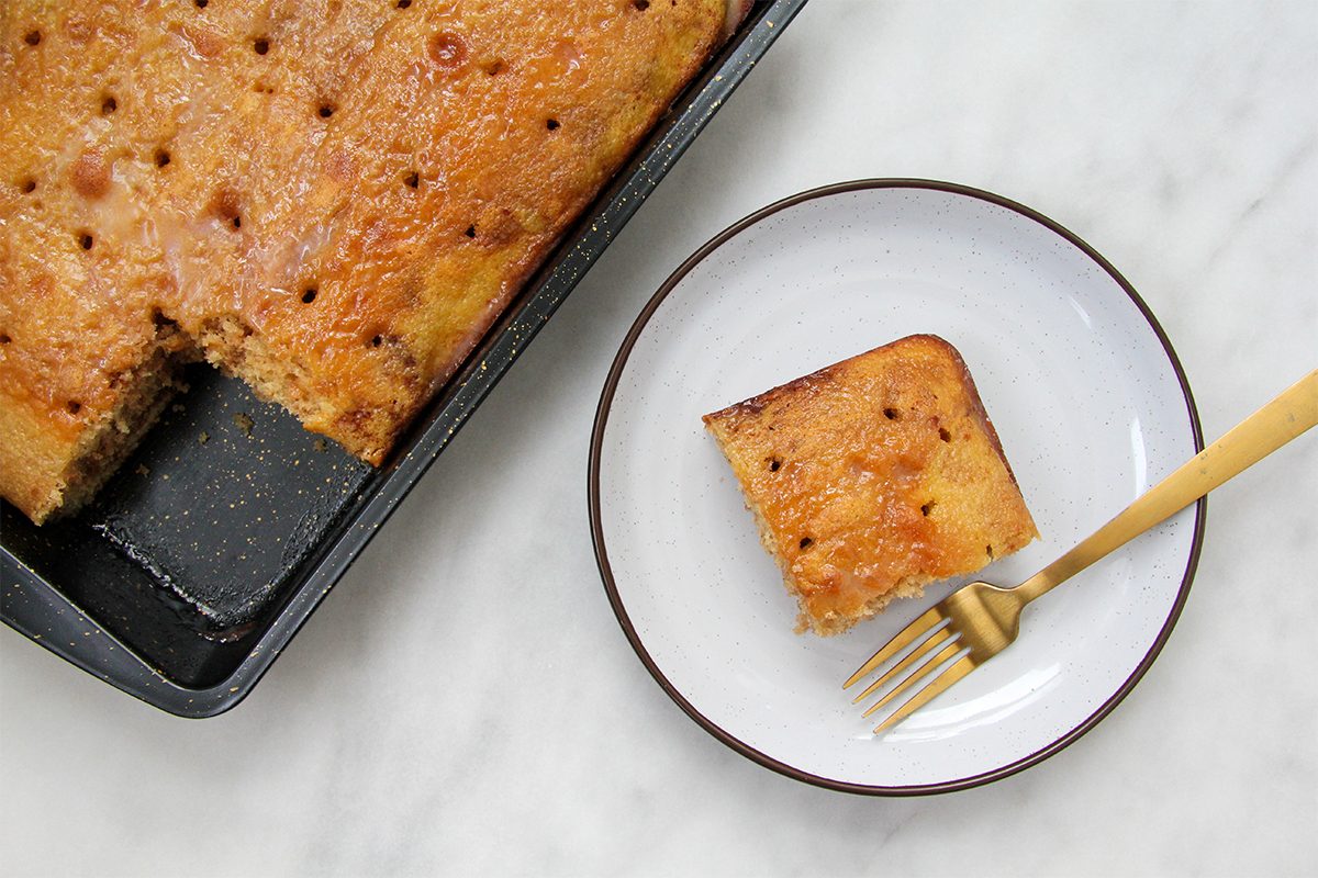 A square slice of cake on a white plate with a black rim, accompanied by a gold fork. The cake is inside a black baking tray with a few pieces missing, and the surface shows small holes. The background is a white marble surface.
