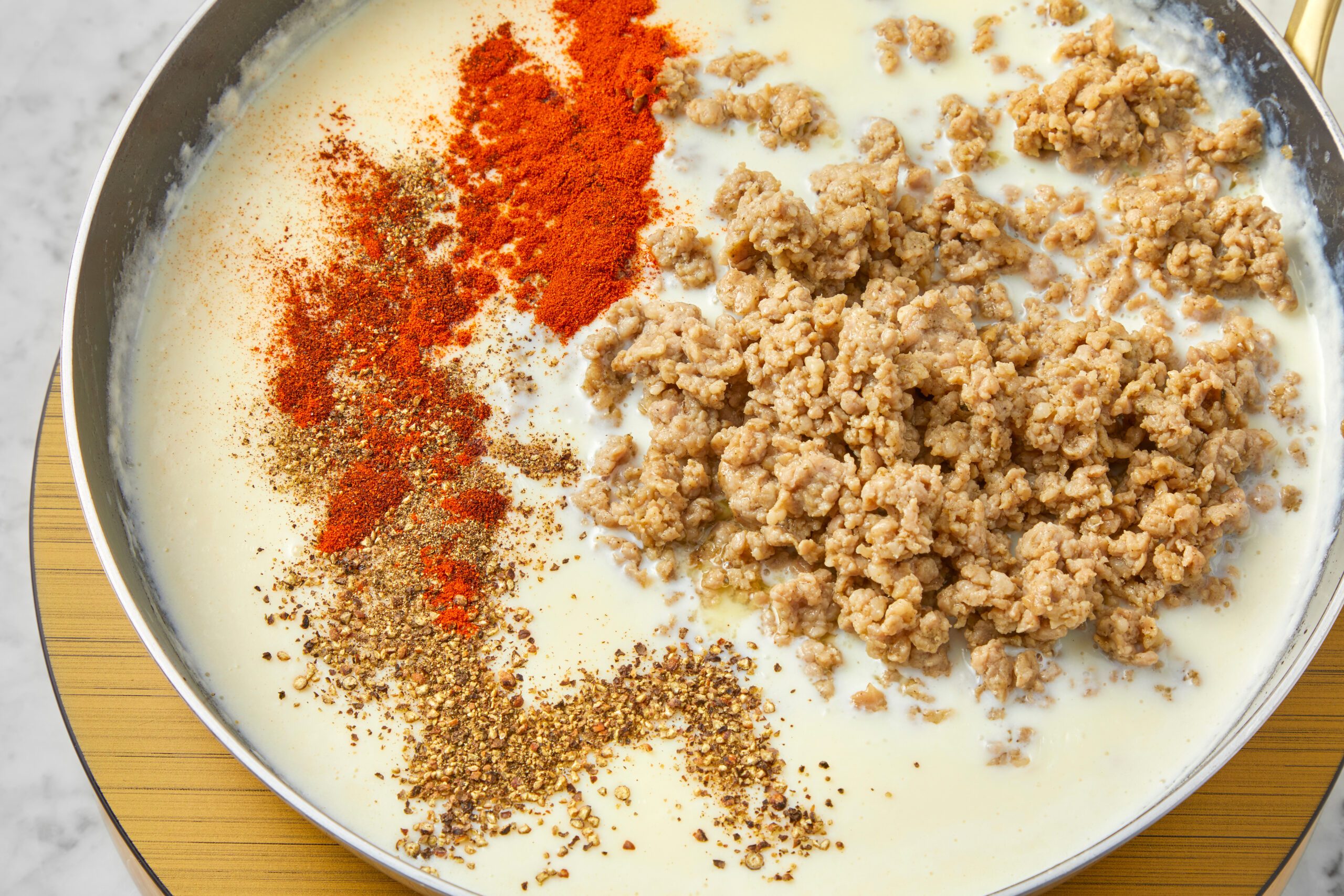 A skillet filled with a creamy white sauce, topped with ground meat, red paprika, and brown spices. The ingredients are partially mixed with a visible swirl pattern.