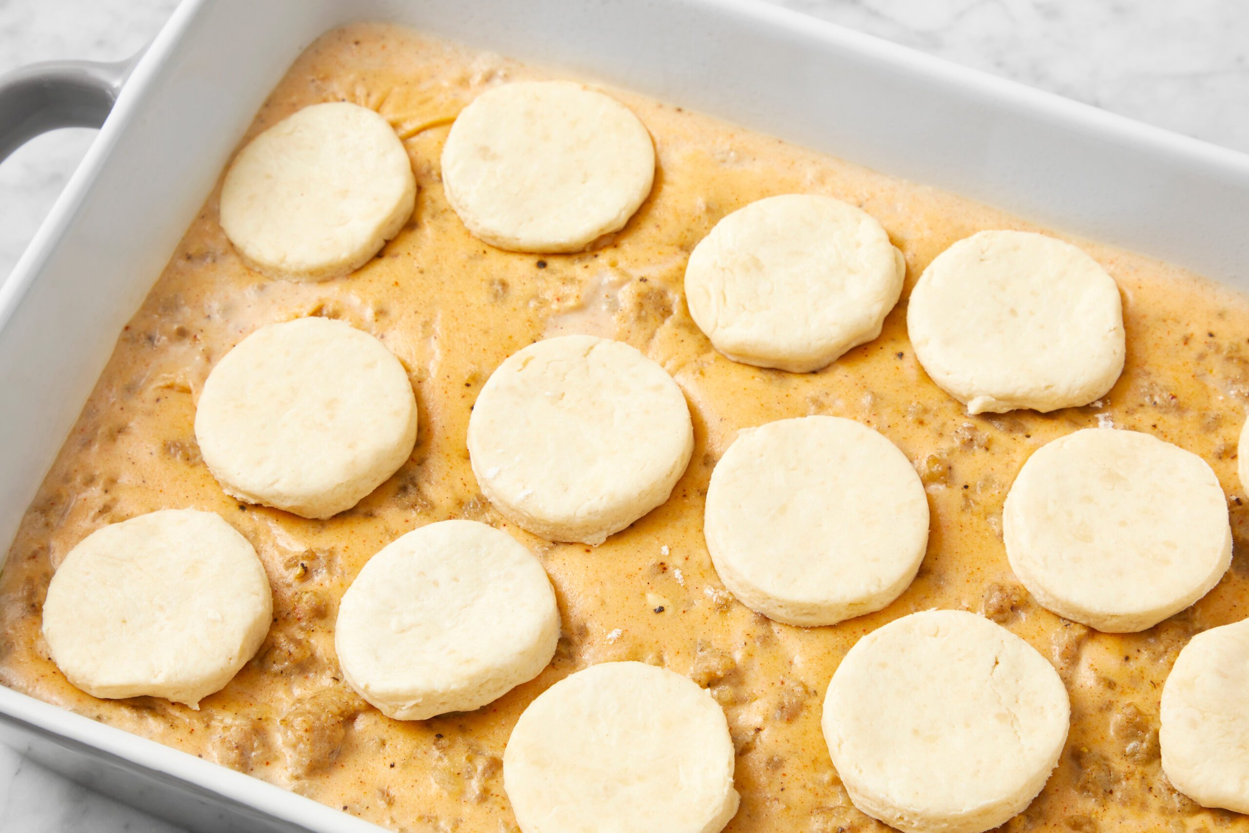 Unbaked biscuits placed evenly over a creamy, orange-colored gravy with visible ground meat, in a rectangular baking dish. The dish is set on a marbled countertop, ready to be baked.