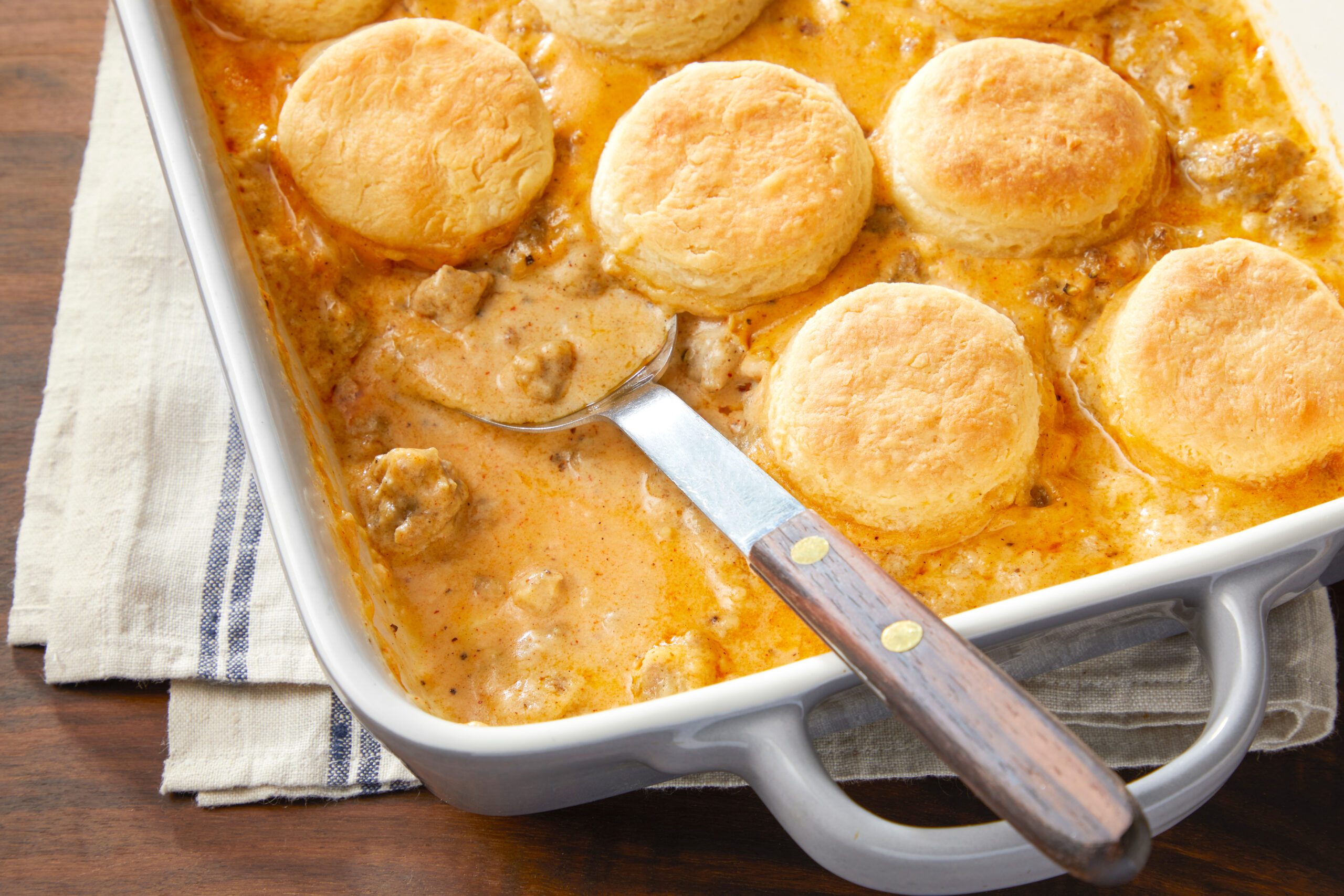 A casserole dish filled with golden-brown biscuits on top of a creamy, sausage gravy mixture. A serving spoon rests in the dish, which is on a striped cloth over a wooden table.
