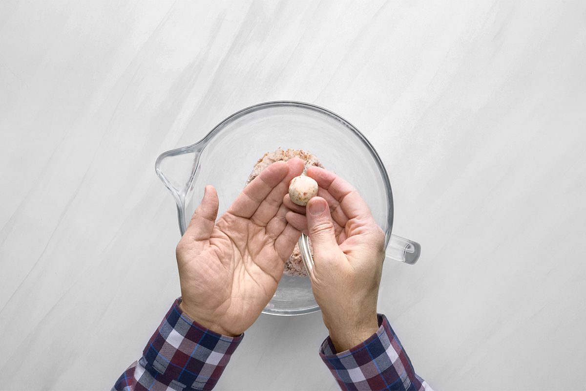 Hands holding an egg over a glass mixing bowl, ready to crack it. The person is wearing a plaid shirt, and the bowl is set on a white countertop.