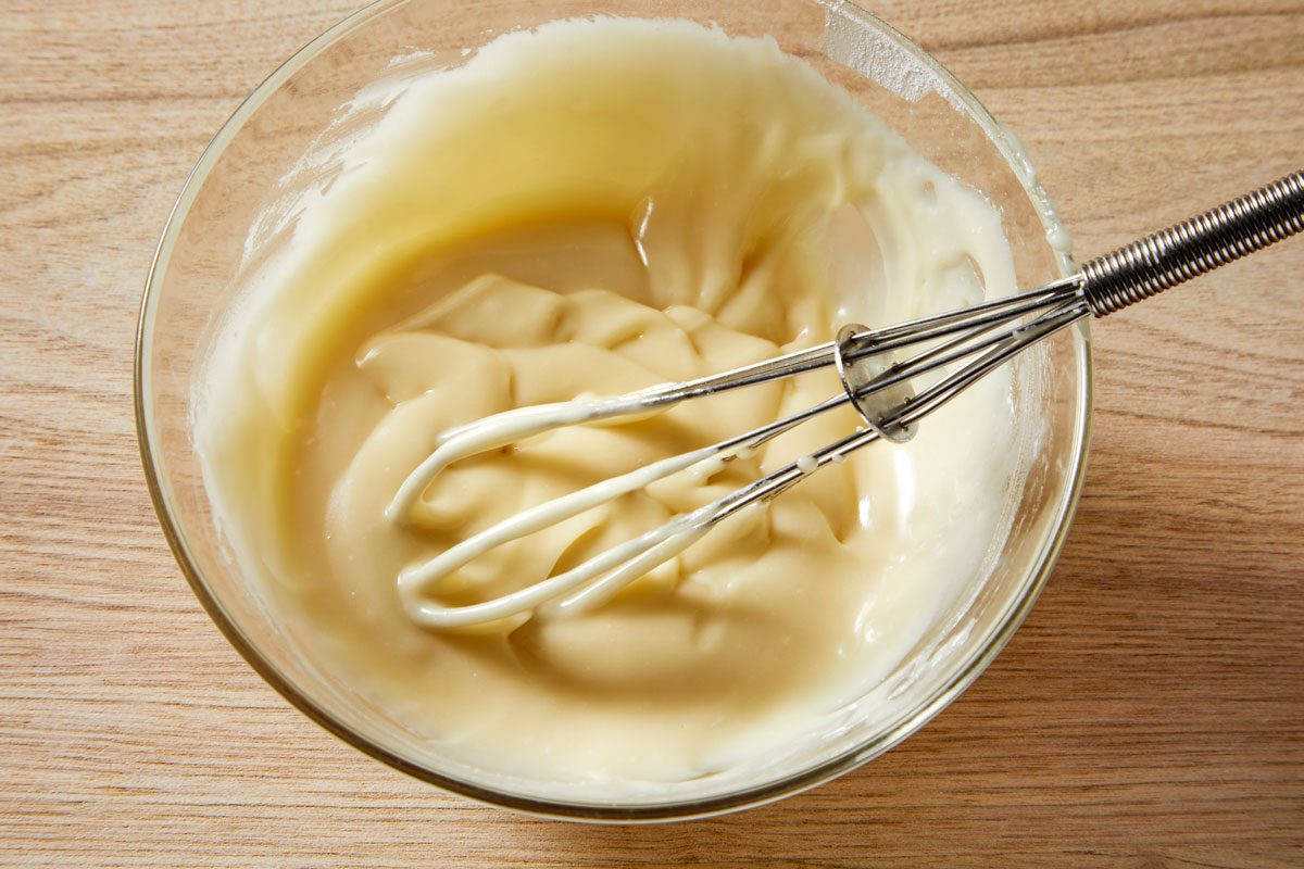 3/4th shot of a glass bowl filled with a light yellow, creamy substance, likely batter or frosting, on a wooden surface, a wire whisk sits in the bowl with the handle extending to the right of the frame