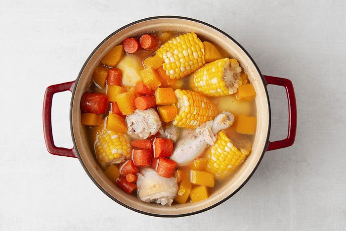 A red-handled pot filled with a stew, containing chicken pieces, corn on the cob, carrots, and chunks of squash in a broth. The ingredients are mixed together and visible from a top angle.