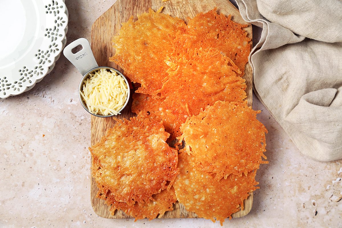 A wooden board with several crispy cheese crisps and a small metal cup filled with shredded cheese. A beige cloth lies beside the board, and a decorative white plate is partially visible in the top left corner.