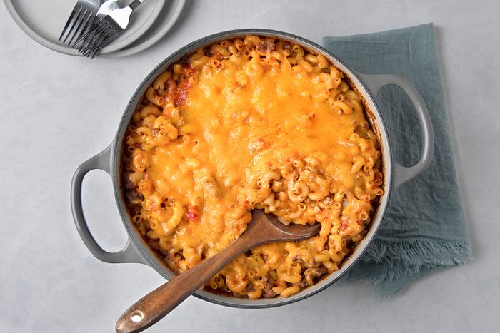 A pot of baked macaroni and cheese with a golden-brown crust. A wooden spoon is resting in the dish, and a green cloth napkin and a stack of plates with forks are nearby on a light gray surface.