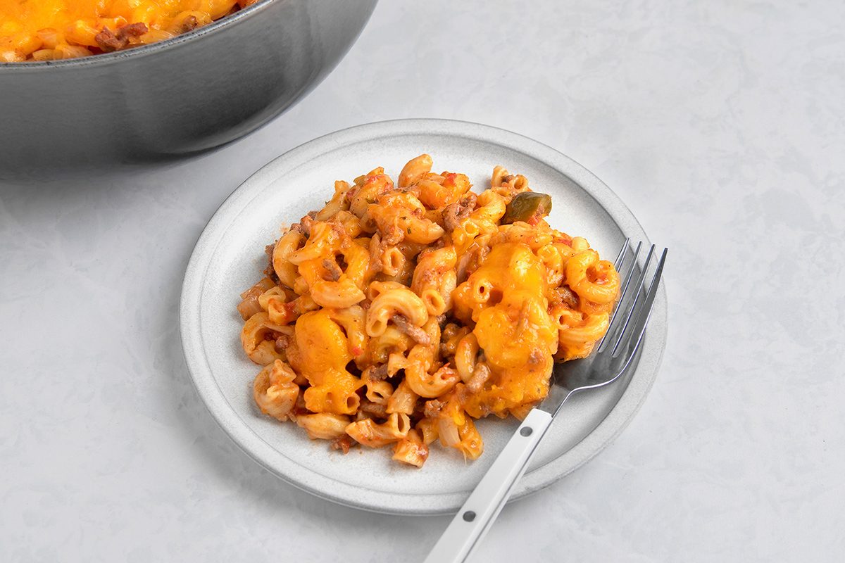 A white plate with baked mac and cheese, featuring elbow macaroni and melted cheddar cheese. A fork rests on the plate, and a portion of the dish is visible in a pot in the background, all on a light countertop.