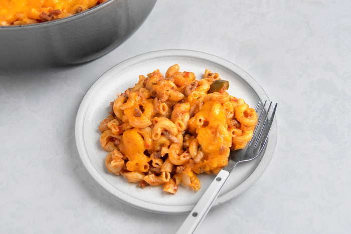 A white plate with baked mac and cheese, featuring elbow macaroni and melted cheddar cheese. A fork rests on the plate, and a portion of the dish is visible in a pot in the background, all on a light countertop.