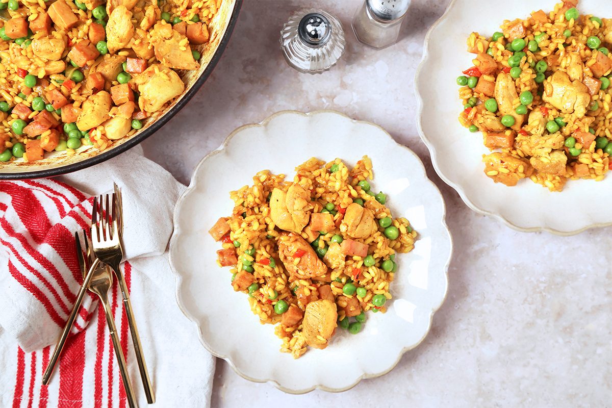 Plates of chicken fried rice with peas and carrots are arranged on a light countertop. A skillet with more of the dish and a red-striped napkin with forks are nearby, along with salt and pepper shakers.