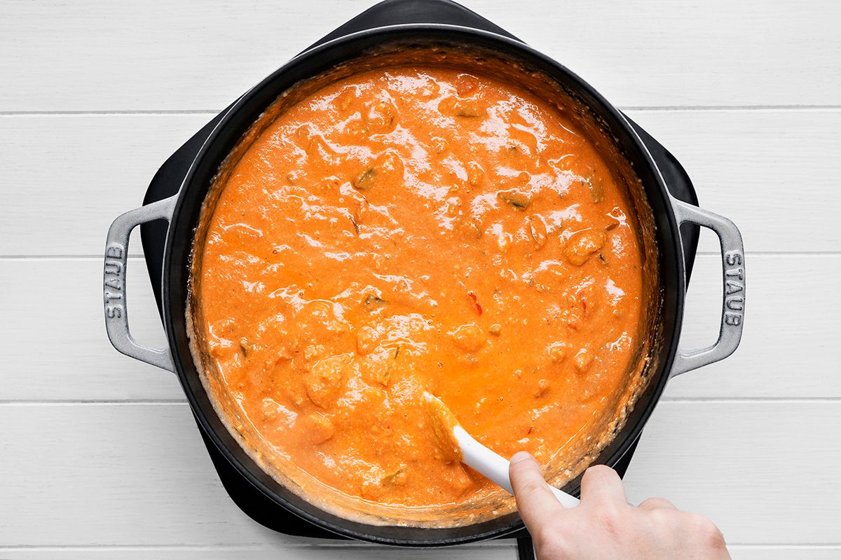 A hand stirring a pot of creamy orange tomato soup with a white spoon. The pot is a black Staub, and it sits on a wooden surface. The soup appears to have small chunks, likely ingredients, blended into the smooth consistency.