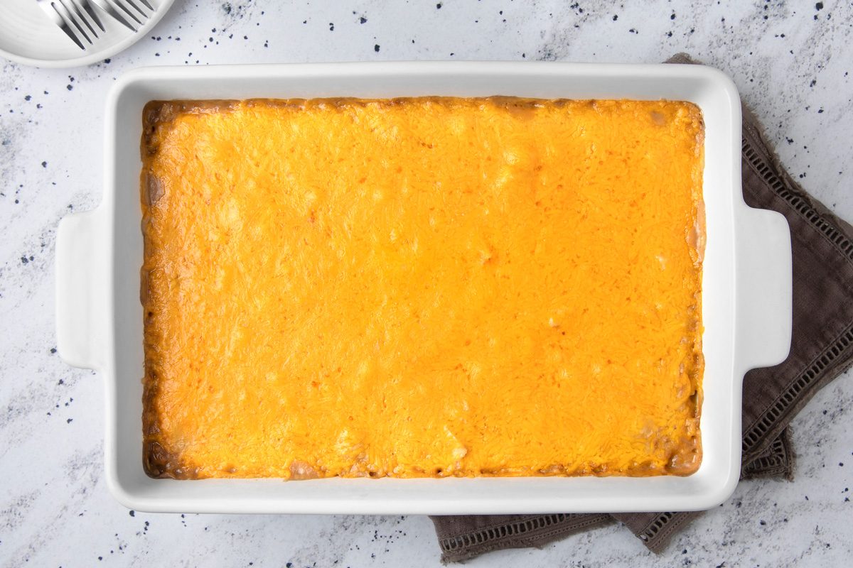 overhead shot of a white baking dish filled with a golden-yellow cheesy casserole; The baking dish is placed on a white surface with a light brown cloth napkin beside it, a white plate with silver forks lying in the top left corner of the image