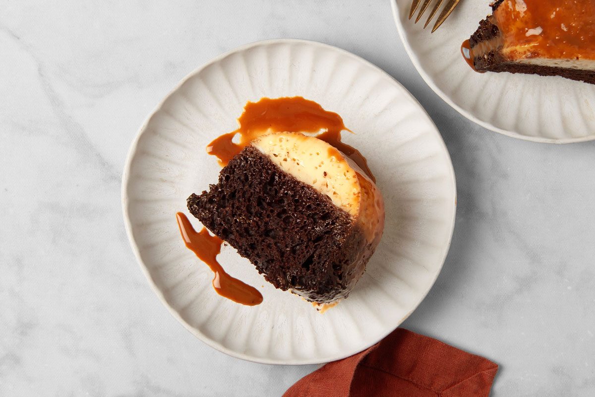 overhead shot of a slice of Chocoflan, resting on a white plate with a rippled design, which is set on a marble countertop; a partially visible orange napkin sits at the bottom of the frame, In the background, another white plate with a fork is visible;