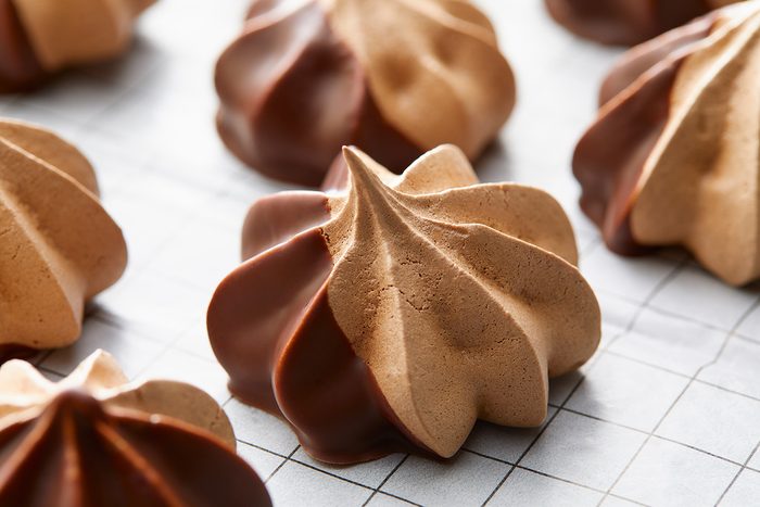 Brown meringue cookies with a glossy chocolate-dipped base are arranged on a white grid-patterned surface. The cookies have a swirled, pointed shape, showcasing a contrast between the matte meringue and shiny chocolate.