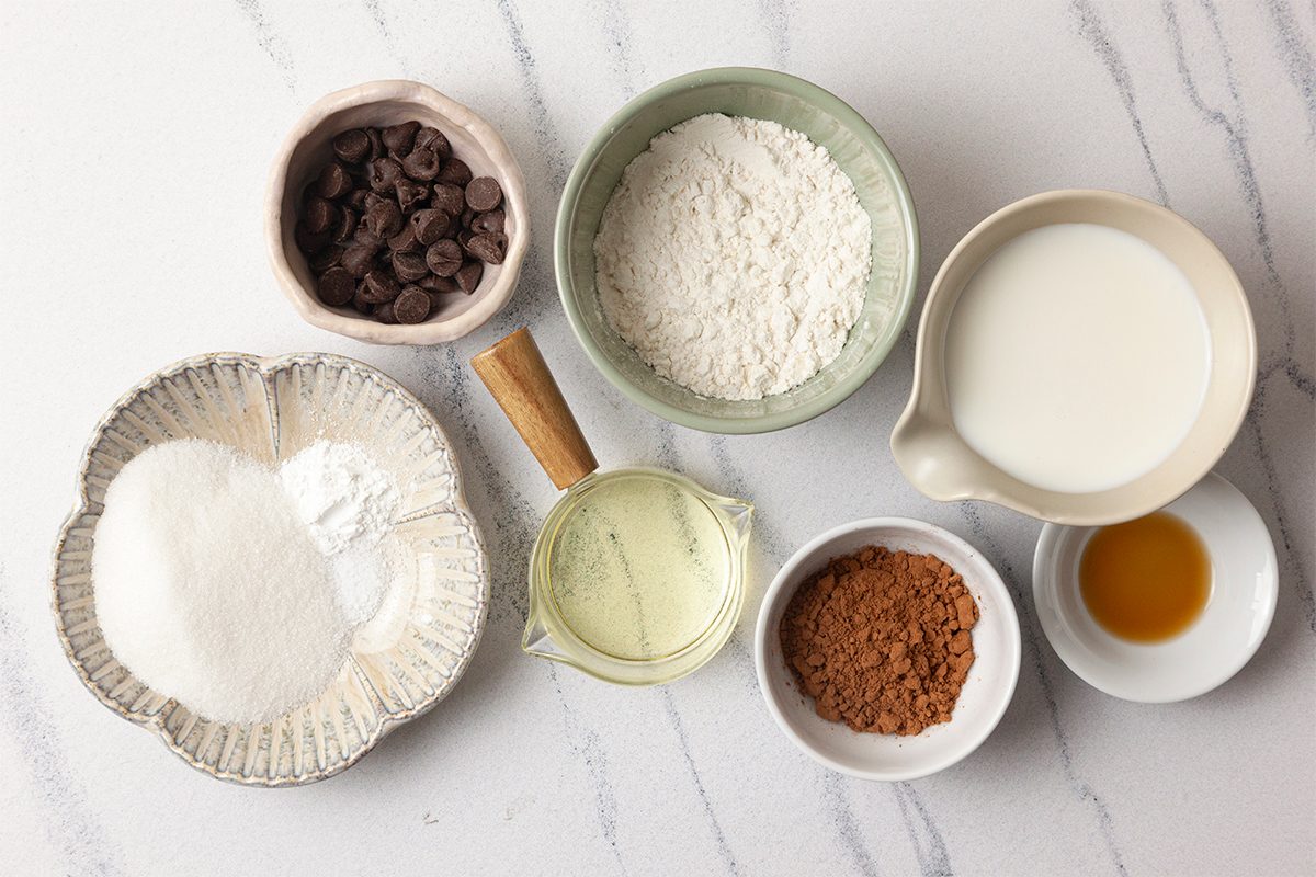 Various baking ingredients arranged on a marble surface: chocolate chips, flour, milk, sugar, baking powder, cocoa powder, vegetable oil, and vanilla extract in separate bowls and a measuring cup.