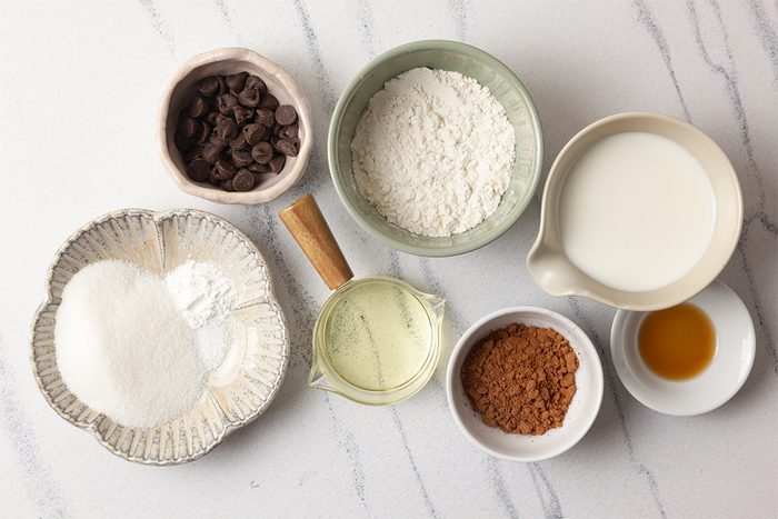 Various baking ingredients arranged on a marble surface: chocolate chips, flour, milk, sugar, baking powder, cocoa powder, vegetable oil, and vanilla extract in separate bowls and a measuring cup.