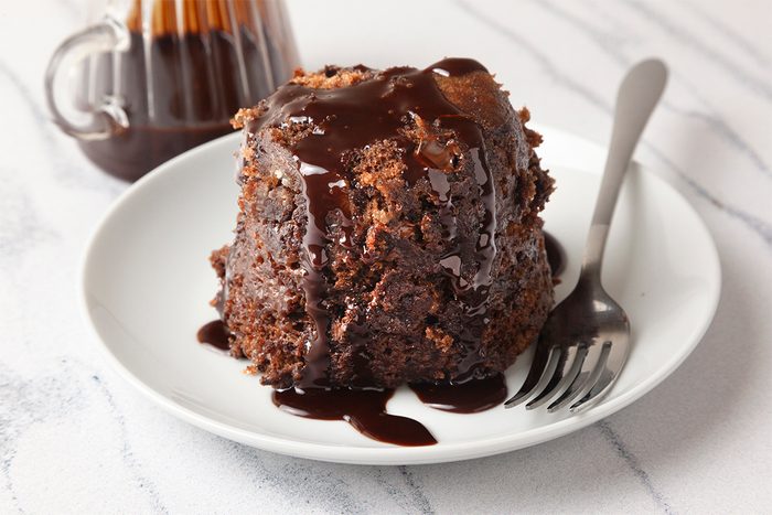 A chocolate lava cake sits on a white plate, topped with rich chocolate sauce that drips down the sides. A fork rests beside it. A glass jug filled with additional chocolate sauce is in the background on a marble surface.