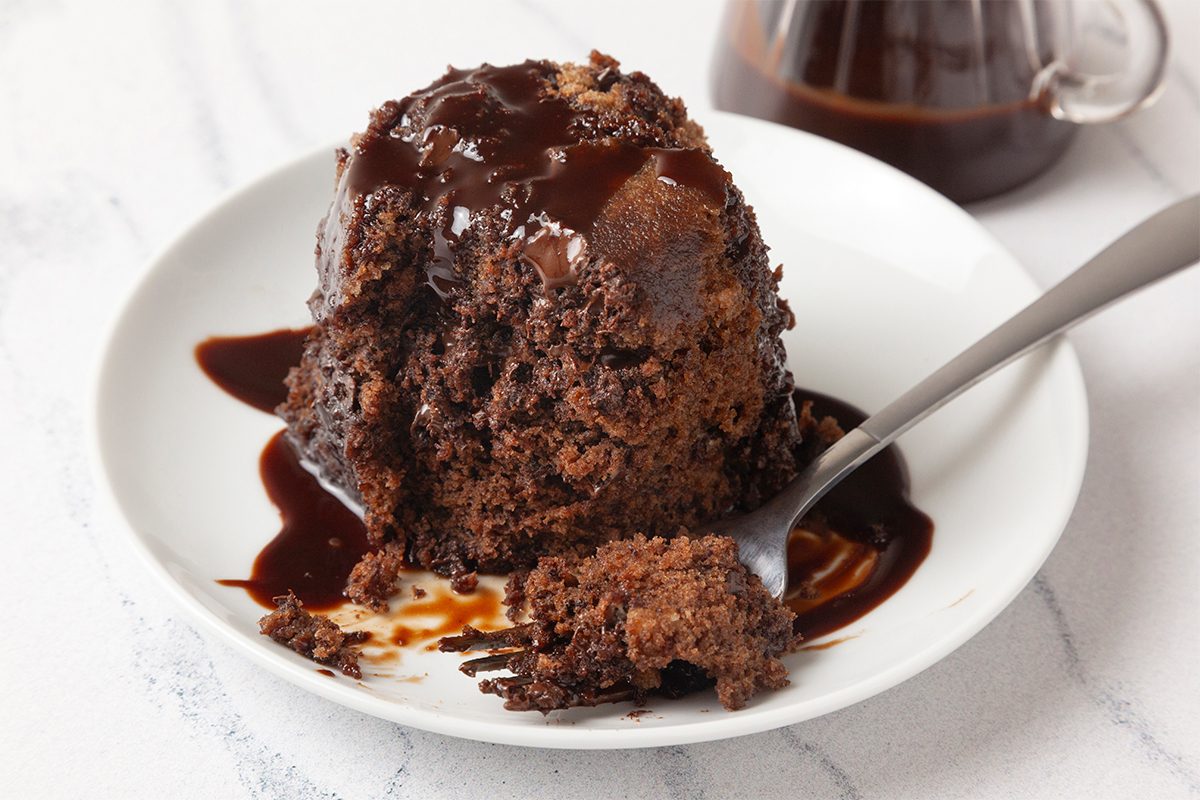 A rich chocolate cake with a molten center is served on a white plate. It is generously drizzled with chocolate sauce, and a spoon is resting beside it, ready to take a bite. A jar of chocolate sauce is in the background.