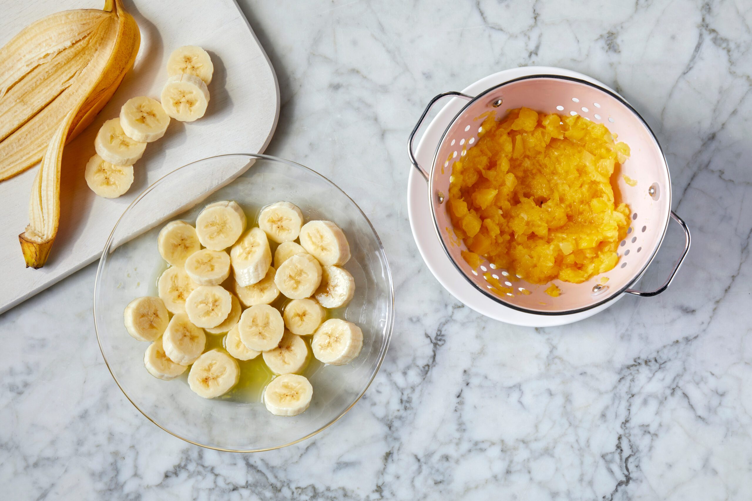 Sliced bananas on a plate, a peeled banana on a cutting board, and crushed pineapple in a colander on a marble countertop.