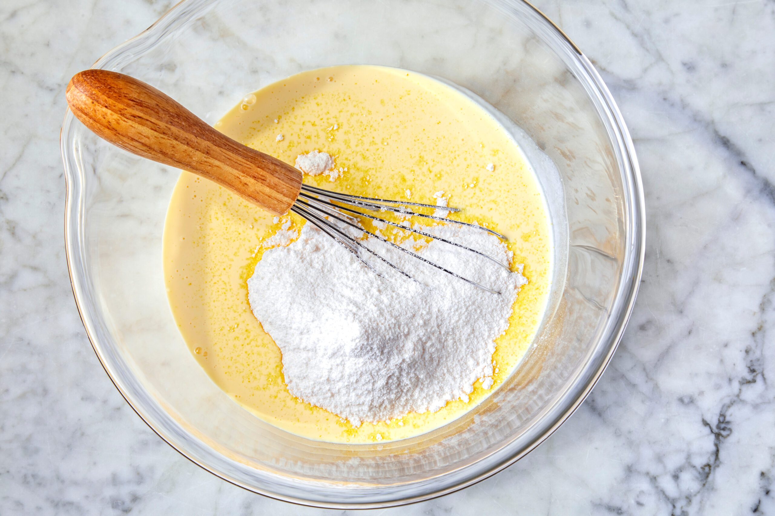 A glass bowl on a marble countertop contains a yellow liquid mixture with a wooden-handled whisk and a mound of white flour on top.