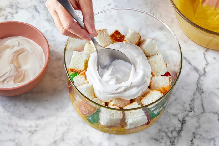 A hand holding a spoon is spreading whipped cream over a layered dessert in a glass bowl. The dessert includes pieces of white cake and colorful gelatin. A bowl with more whipped cream is on the side. The scene is set on a marble countertop.