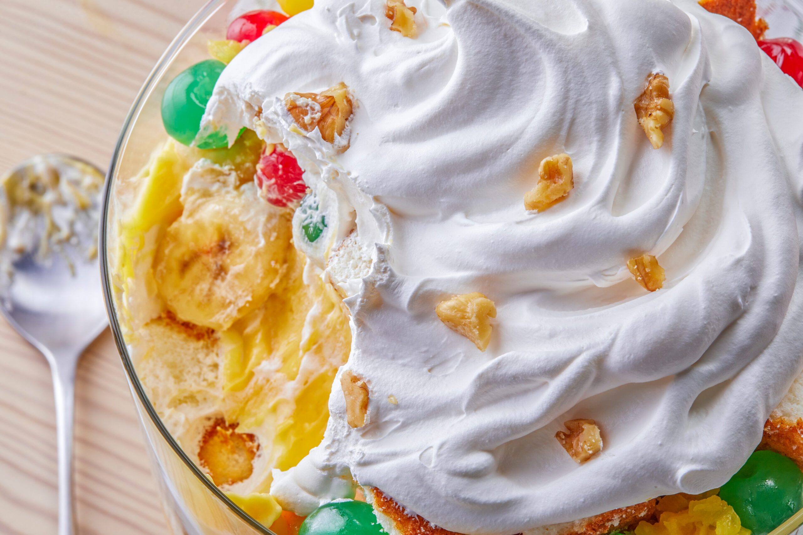A close-up of a dessert featuring layers of whipped cream, colorful jelly candies, and crushed nuts on top. A spoon with remnants of the dessert is visible to the left on a wooden surface.