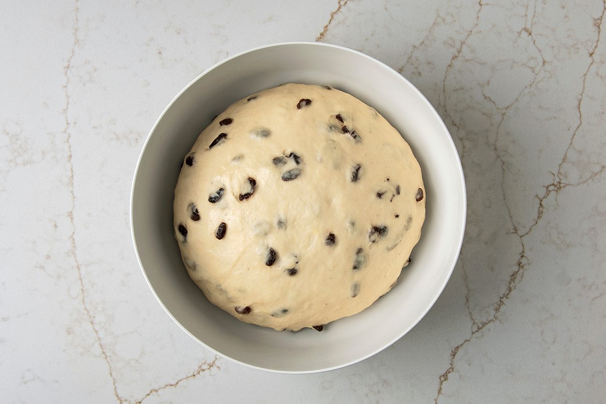 A ball of dough with raisins is set in a white mixing bowl, placed on a marbled countertop.