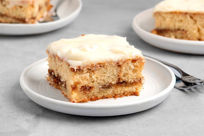 A slice of layered cake with a white frosting on top, served on a white plate. The cake has a light brown color and appears moist, with another similar slice visible in the background on a separate plate. Silver forks accompany each plate.