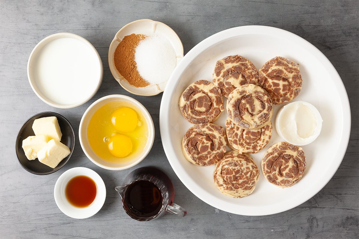 A flat lay of ingredients for cinnamon rolls. A large plate with unbaked rolls, bowls containing eggs, butter, cream, sugar, brown sugar, vanilla extract, and syrup are arranged on a gray surface.
