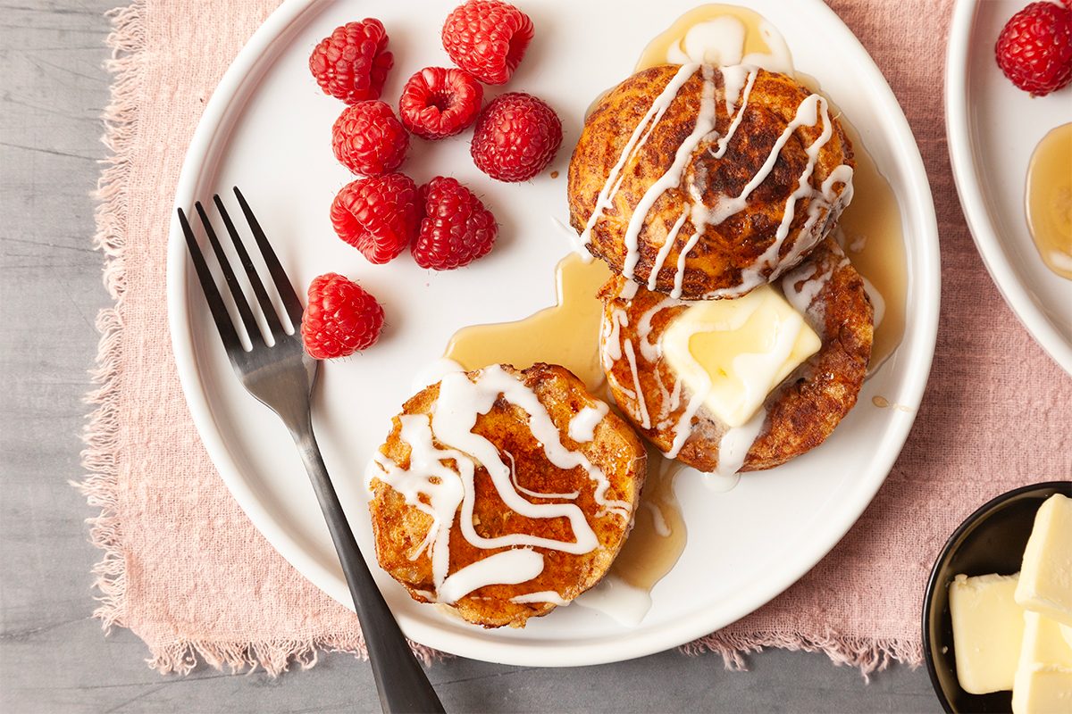 A white plate with two pancakes topped with a pat of butter and white drizzle. Syrup is poured over them. Fresh raspberries are on the side. A fork rests on the plate. A small dish with more butter is nearby, all on a pink cloth.