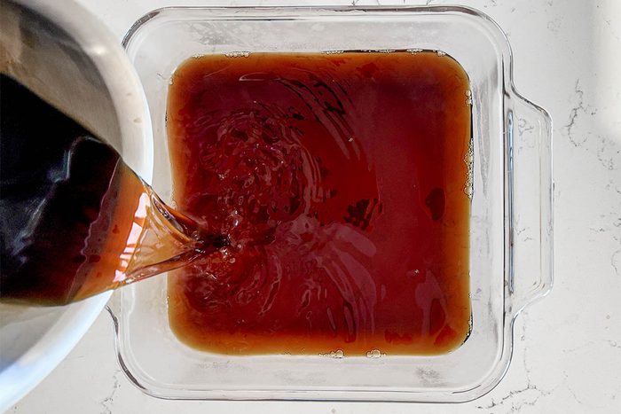 A dark liquid is being poured from a pot into a clear, square container on a white countertop. The liquid is rich brown, creating swirls as it fills the container.