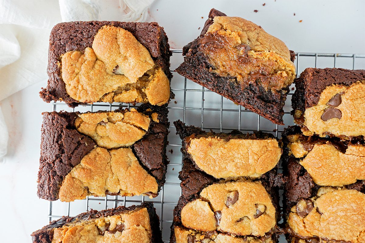 Brownies with chocolate chip cookie tops arranged on a cooling rack. Some brownies are cut into squares, showing a contrast between the dark chocolate base and golden cookie layer. A white cloth is partially visible in the background.