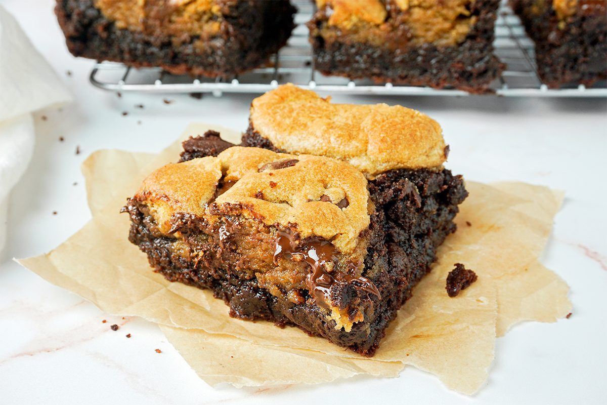 A close-up of a fudgy brownie topped with golden, toasted marshmallows. It's on a piece of parchment paper, with a cooling rack and more brownies in the background. The brownie appears rich and gooey.