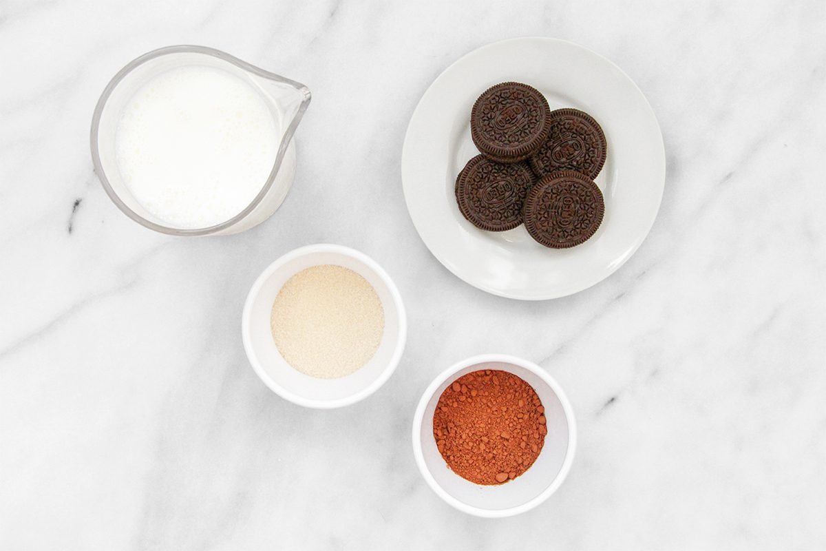 A marble countertop with a small pitcher of milk, a plate of chocolate sandwich cookies, and two bowls: one with white sugar and another with reddish-brown sugar.