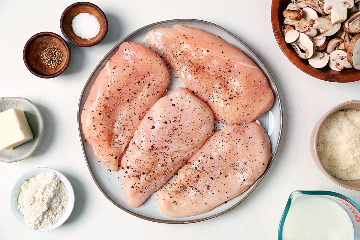 A plate with raw chicken breasts seasoned with pepper. Surrounding the plate are bowls of mushrooms, salt, pepper, grated cheese, and flour. There is also a piece of butter and a glass container with milk.